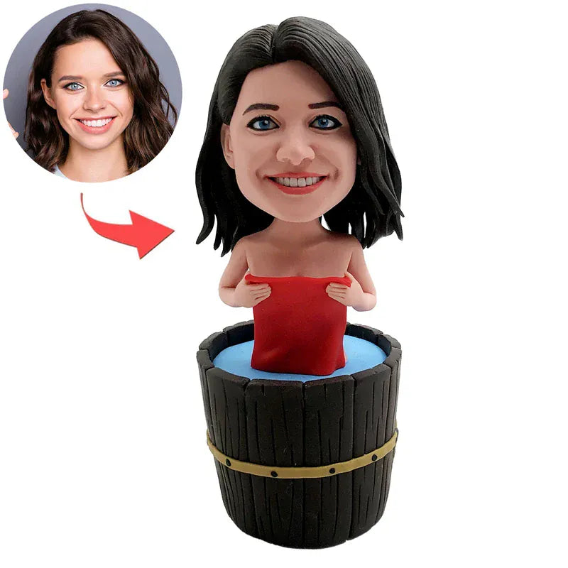 Custom female bobblehead relaxing in a wooden bathtub with a red towel, displayed on a spa-style shelf with rolled towels, soap, and bath salts.webp