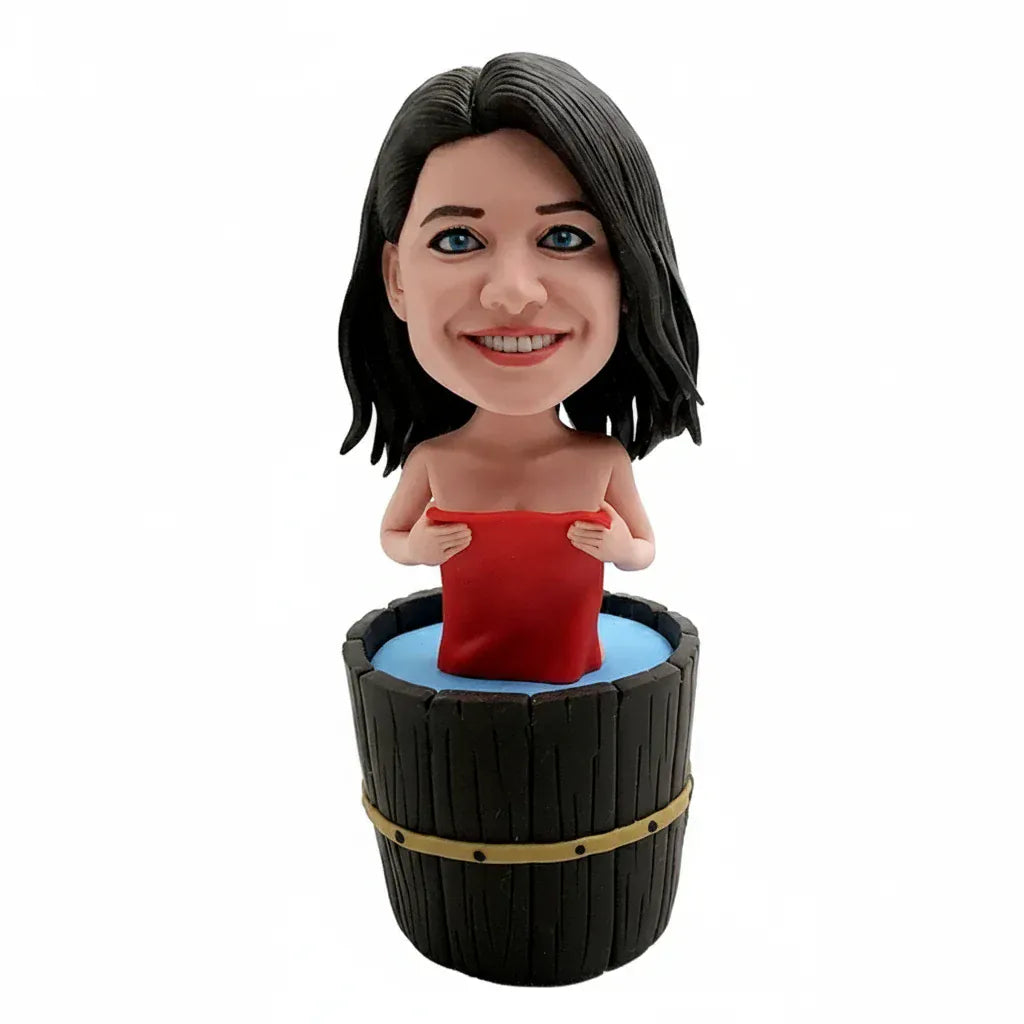 Custom female bobblehead relaxing in a wooden bathtub with a red towel, displayed on a spa-style shelf with rolled towels, soap, and bath salts